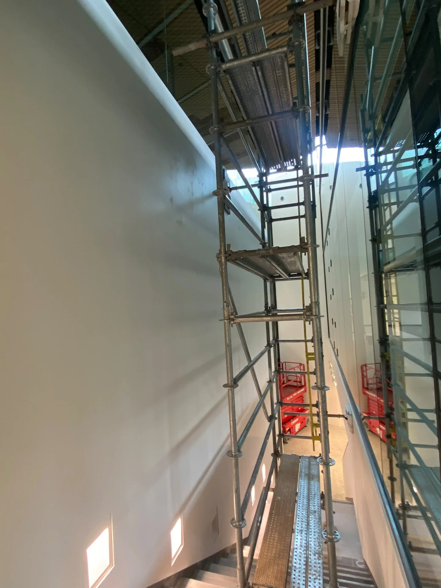 Scaffolding beside a tall light-grey wall beneath an exposed commercial ceiling